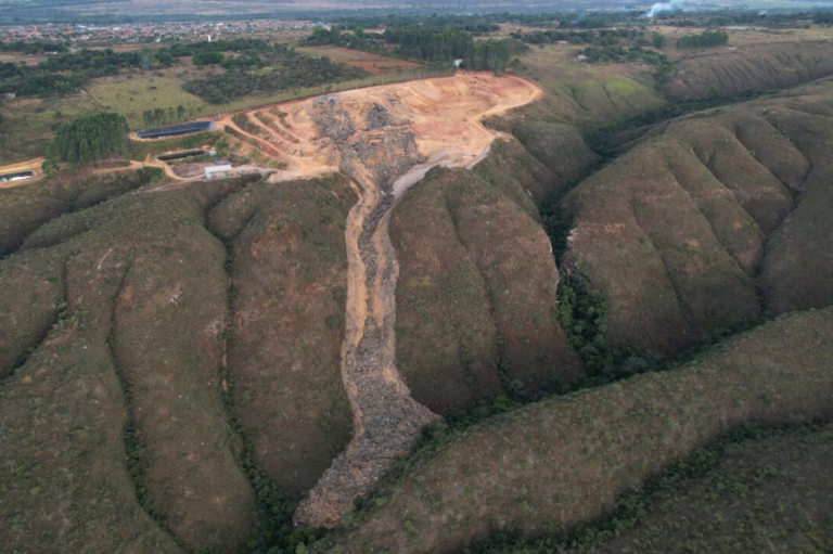 A imagem mostra uma paisagem natural vista de cima (aérea), com destaque para uma área de desmatamento e intervenção humana em meio a formações montanhosas. No centro da imagem, observa-se uma clareira extensa com solo exposto e marcas de movimentação de terra, indicando atividade de mineração ou terraplanagem. A terra removida escorre em direção a um vale estreito entre as colinas, formando uma espécie de cicatriz no relevo. A vegetação nativa, que cobre as encostas arredondadas das colinas, foi retirada dessa área, evidenciando o impacto ambiental da atividade. Ao fundo, é possível ver áreas verdes, plantações, construções dispersas e uma cidade mais afastada, sugerindo a proximidade entre zonas urbanas e a área degradada.