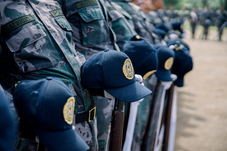 Foto mostra detalhe de dezenas de pessoas de pé em formação, lado a lado, com uniformes da polícia, tendo à frente uma espingarda cada um e um boné colocado sobre a ponta da arma. A imagem não mostra rostos.