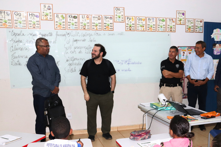 Foto em ambiente de sala de aula, onde um grupo de adultos, incluindo o procurador da República Sérgio Cipriano ao centro, está de pé em frente a uma lousa branca, conversando com crianças sentadas em carteiras escolares em primeiro plano.