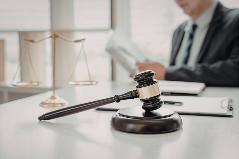 A imagem apresenta uma composição clássica e profissional que remete ao universo jurídico e à justiça. Abaixo, detalho os principais elementos da cena:

Elementos em Primeiro Plano
Martelo (Gavel): Um martelo de madeira escura com detalhes em metal dourado repousa sobre sua base circular. Ele é o ponto focal da imagem, simbolizando autoridade, ordem e o veredito final.

Escritório: A superfície da mesa é branca e limpa, sugerindo um ambiente organizado e sério.

Elementos em Segundo Plano
Balança da Justiça: À esquerda, observa-se uma balança dourada, símbolo universal da equidade e do equilíbrio nas decisões judiciais.

Figura Humana: Ao fundo, de forma desfocada (efeito bokeh), vê-se um homem vestindo um traje formal (paletó escuro, camisa clara e gravata). Ele parece estar manuseando documentos ou lendo um processo, reforçando a ideia de trabalho jurídico diligente.

Papelada: Sobre a mesa, há uma prancheta com papéis e uma caneta, indicando a preparação de documentos legais.