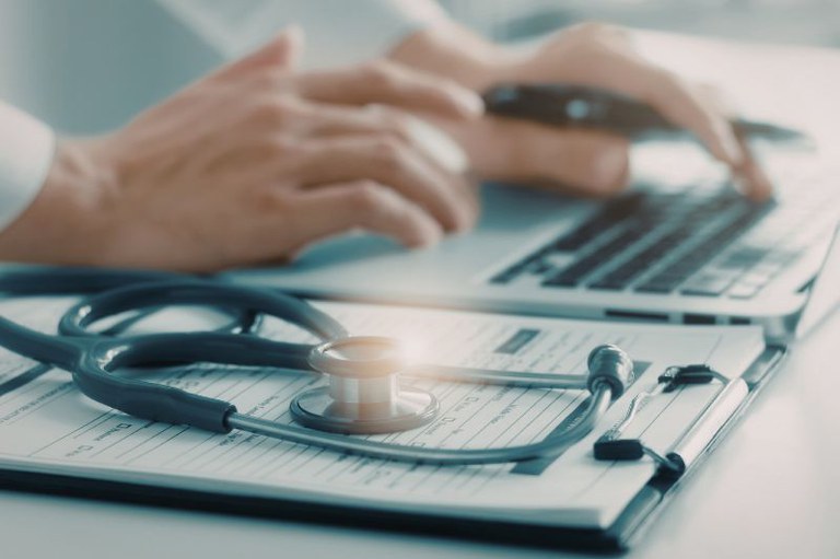 Imagem de uma pessoa no computador e um estetoscópio sobre uma prancheta ao lado