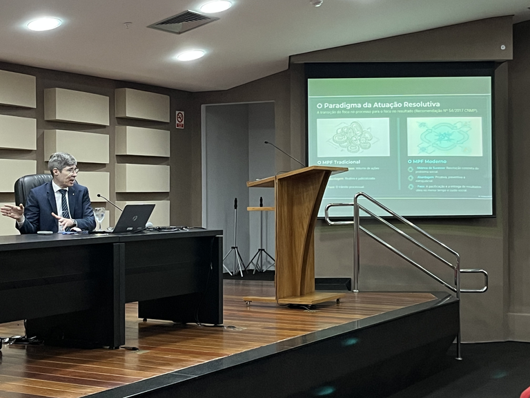 Procurador Marcello Wolff, de cabelos grisalhos, sentado atrás de uma mesa em um auditório, fala enquanto gesticula com uma das mãos. Ele usa terno escuro, camisa social e gravata, e está voltado para a esquerda da imagem. Sobre a mesa há um notebook aberto, um copo de água e outros objetos.

À frente, há um púlpito de madeira sobre um pequeno palco com piso de madeira. Ao fundo, um telão exibe um slide com título e conteúdos em texto e imagens, não totalmente legíveis.

O ambiente é interno, com iluminação artificial no teto e paredes com painéis acústicos. À direita, há uma rampa com corrimão que dá acesso ao palco. Não há outras pessoas visíveis na imagem.