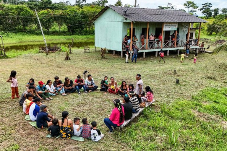Há um grupo de aproximadamente 20 pessoas sentadas em círculo sobre a grama. O grupo é misto, composto por adultos, jovens e crianças. Ao fundo, vê-se uma construção de madeira elevada do chão (tipo palafita), pintada de verde-claro.
