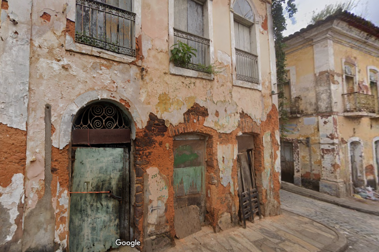 Foto mostra fachada de imóvel com pintura descascada, pedaços de madeira no lugar de portas e janelas, em estado precário