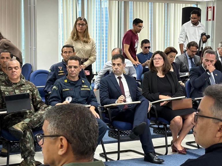 Participantes acompanham a solenidade de instalação do Comitê de Segurança das Eleições 2026, realizada em uma sala de reuniões do Tribunal Regional Eleitoral do Maranhão (TRE-MA). Em primeiro plano, autoridades civis e militares estão sentadas em cadeiras azuis, entre elas o procurador-chefe da República no Maranhão, Alexandre Soares, de terno escuro e gravata vinho, além de representantes da Marinha do Brasil, do Exército Brasileiro e de outros órgãos. Ao fundo, jornalistas e servidores registram e acompanham o evento. O ambiente possui amplas janelas com persianas verticais e disposição típica de auditório.