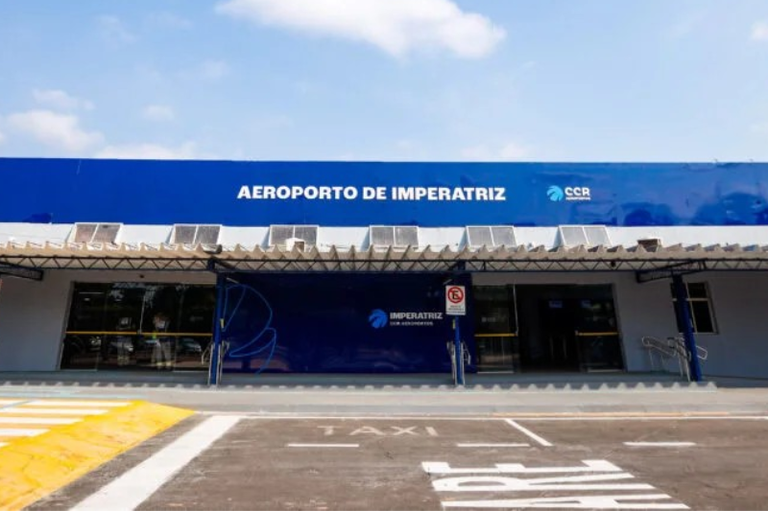 Fachada do Aeroporto de Imperatriz em dia ensolarado. O prédio tem estrutura baixa, com faixa azul na parte superior e o nome "Aeroporto de Imperatriz" em destaque. À frente, há área de embarque e desembarque, faixa de pedestres e sinalização para táxi. O céu está azul, com poucas nuvens.