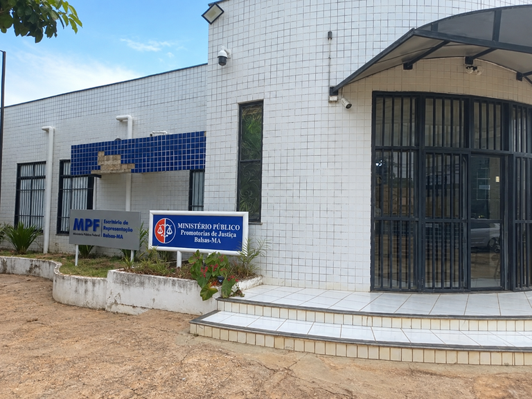 Fotografia colorida da fachada de um prédio público administrativo em um dia claro, sob um céu azul com nuvens esparsas. A construção possui paredes revestidas por pequenos azulejos brancos quadrados e apresenta uma estrutura de formato levemente arredondado do lado direito, onde fica a entrada principal. O acesso a essa entrada é feito por uma pequena escada de três degraus revestidos de cerâmica branca, que leva a um grande portão protegido por grades de metal preto com linhas verticais. Acima desse portão, há uma cobertura curva ou toldo de cor escura e, fixadas na parede externa, veem-se duas câmeras de segurança brancas em alturas diferentes. No centro da fachada, há uma janela vertical estreita e alta com uma grade decorativa de ferro. Mais à esquerda, a parede recua um pouco, exibindo uma seção decorada com azulejos azuis escuros, onde alguns parecem estar danificados ou faltando, logo acima de outras janelas protegidas por grades pretas. Um canteiro baixo de concreto pintado de branco, contendo terra, plantas verdes e algumas flores vermelhas, contorna essa lateral esquerda do edifício. À frente desse canteiro, estão fixadas duas placas de identificação lado a lado: a da direita é azul e traz o logotipo do Ministério Público com uma balança da justiça estilizada dentro de um círculo vermelho, acompanhada do texto em letras brancas "MINISTÉRIO PÚBLICO - Promotorias de Justiça - Balsas-MA"; a da esquerda é cinza e exibe a sigla "MPF" em azul escuro junto ao texto "Escritório de Representação - Ministério Público Federal - Balsas-MA". O solo em frente ao prédio é de terra batida ou areia em tons de marrom e bege claro, e algumas folhas verdes de uma árvore aparecem no canto superior esquerdo da imagem, fora do enquadramento principal.