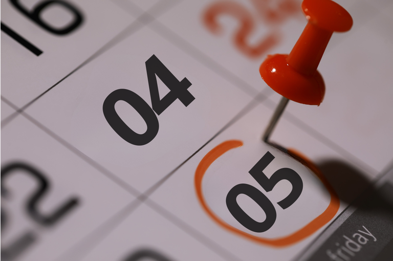 Descrição de acessibilidade: Imagem em close de um calendário com foco nos dias 4 e 5. O número 6 está circulado em laranja e marcado com um alfinete vermelho, simbolizando uma data destacada ou compromisso importante. A fotografia possui efeito desfocado nas bordas, com iluminação suave e tons predominantes em branco, preto e laranja.