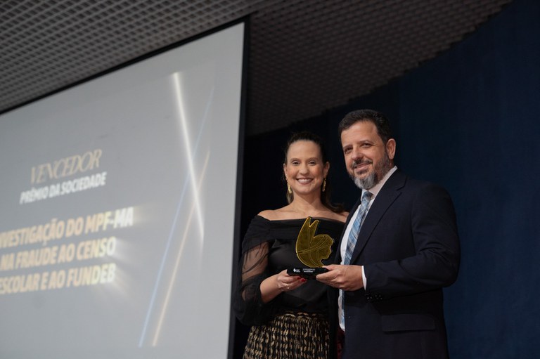 A imagem é uma fotografia do procurador Juraci Guimarães recebendo o troféu do Prêmio Sociedade. O procurador está de terno ao lado de uma mulher de cabelo preso e roupa preta. O prêmio é dourado e se assemelha a um rosto de uma mulher com asas.