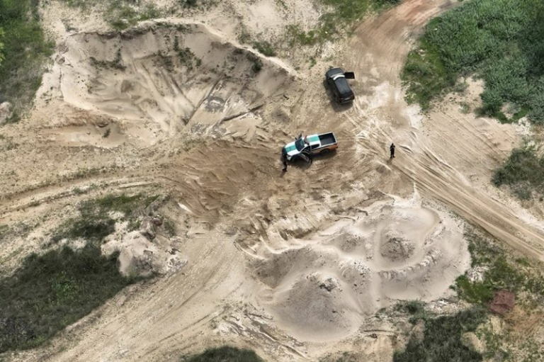 Uma escavadeira hidráulica operando em uma área de extração de areia, com montes de areia e vegetação ao fundo.