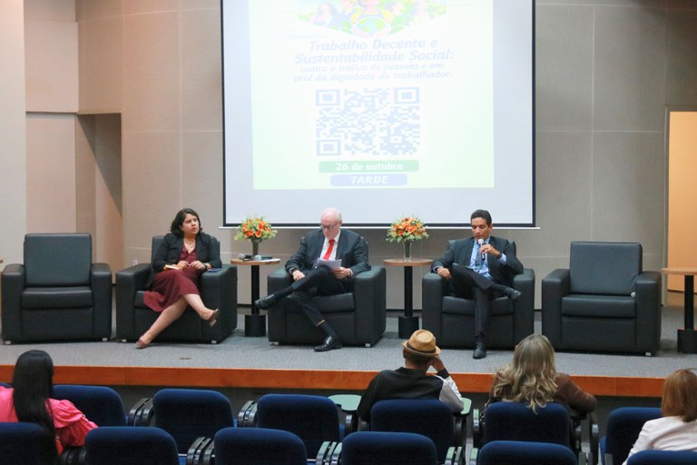 Vista do auditório com três palestrantes dispostos no palco em poltronas escuras. Ao centro, um homem lê papéis, à direita um homem fala ao microfone e à esquerda uma mulher acompanha a apresentação. Ao fundo, uma tela projeta a mensagem "Seminário Trabalho Decente e Sustentabilidade Social: contra o tráfico de pessoas e em prol da dignidade do trabalhador" acompanhada de um QR code. Em primeiro plano, vê-se a plateia de costas.