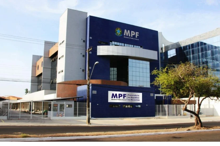 A imagem é uma fotografia da fachada da fachada do prédio do Ministério Público do Estado do Maranhão, com estrutura moderna de vários pavimentos. O edifício apresenta uma fachada predominantemente azul-escura, com detalhes em branco, cinza e marrom, além de janelas amplas de vidro. Na parte superior e na entrada, há placas com a sigla “MPF” e referências à Procuradoria da República no Maranhão. Na frente do prédio, há grades metálicas, portões de acesso, calçada e via pública. À direita da imagem, observa-se uma árvore próxima à calçada.
