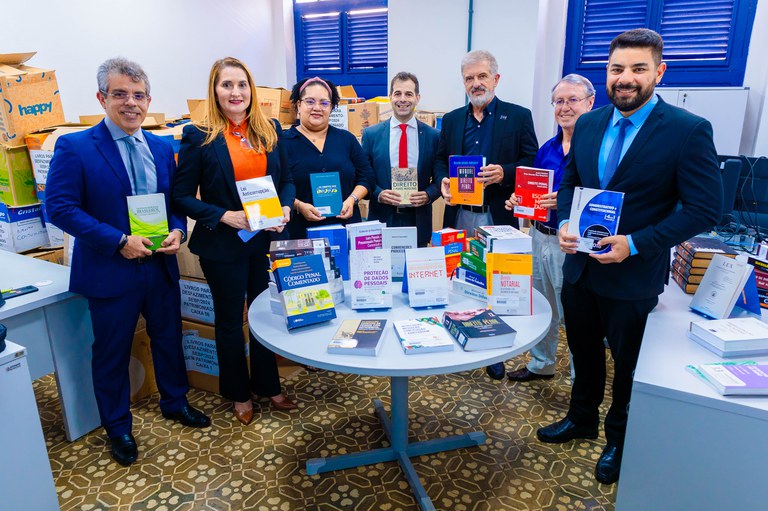 Na imagem, 5 homens e 2 mulheres trajados socialmente com paletas escuras, e nas mãos de cada um está um livro da biblioteca. O ambiente é uma sala fechada, com janelas no fundo, livros em cima de uma mesa. Essas pessoas estão de pé atrás das mesas.