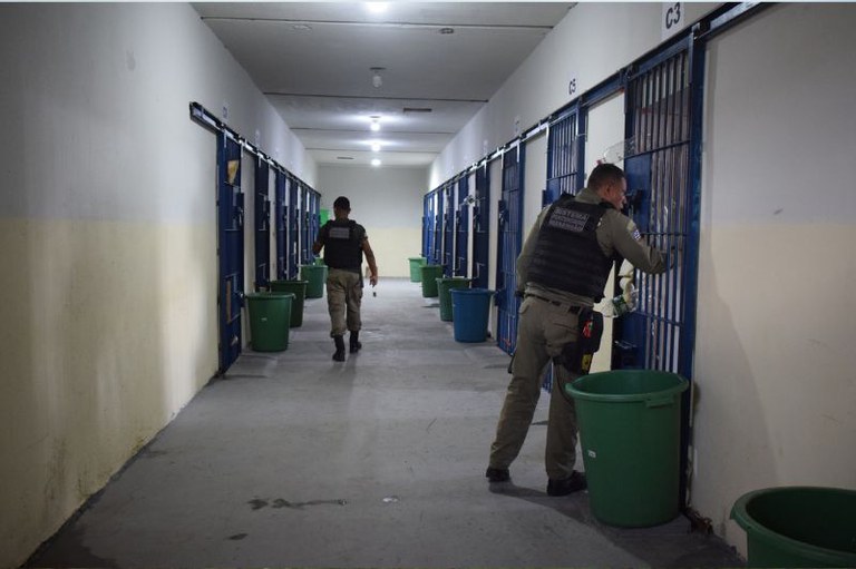 Foto mostra um corredor de uma prisão com celas dos dois lados com portões pintados de azul e dois agentes penitenciários fazendo a fiscalização