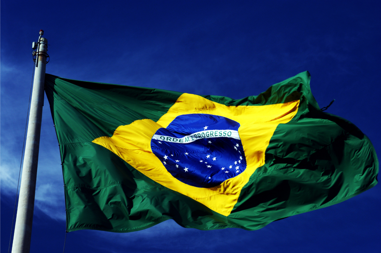Fotografia da bandeira do Brasil hasteada em um mastro, ao ar livre. A bandeira aparece em movimento, com o céu azul ao fundo. No centro, estão o círculo azul com estrelas brancas e a faixa com a inscrição “Ordem e Progresso”, sobre o losango amarelo e o fundo verde.