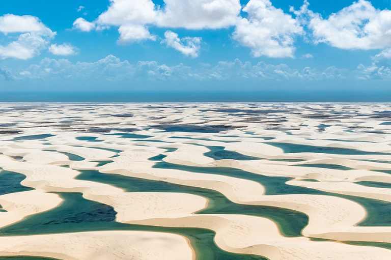 Foto aérea do Parque Nacional dos Lençóis Maranhenses em que se vê diversas dunas de areia clara e lagoas de água entre elas, em dia de céu azul com nuvens