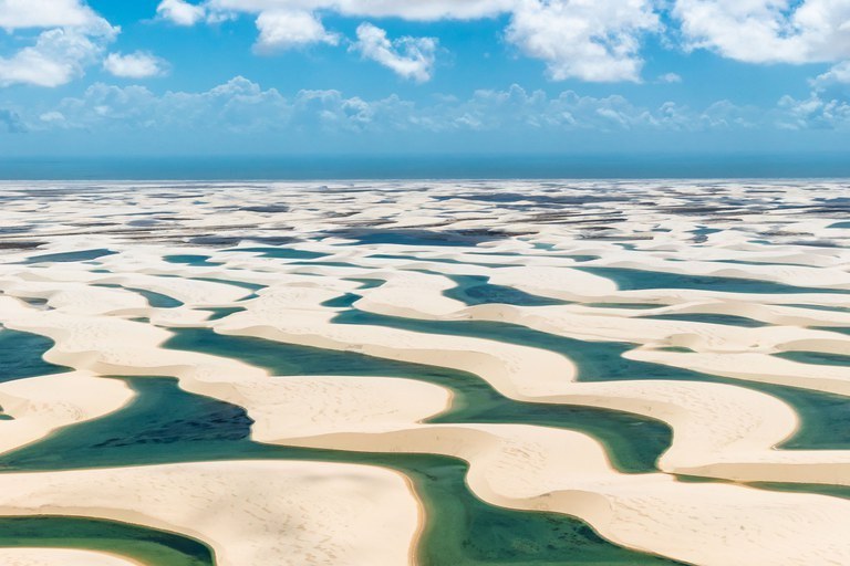 A imagem apresenta uma vista aérea panorâmica do Parque Nacional dos Lençóis Maranhenses, revelando um vasto e hipnotizante deserto de dunas de areia branca e fina que se estende até a linha do horizonte sob um céu azul pontuado por nuvens esparsas. Entre as ondulações das dunas, formam-se inúmeras lagoas de águas cristalinas em tons que variam do verde-esmeralda ao azul-turquesa, criando um contraste vibrante com a aridez do terreno. Ao fundo, a transição suave entre as dunas e o oceano sugere a imensidão costeira dessa paisagem natural única, onde o vento e a chuva moldam constantemente o cenário.