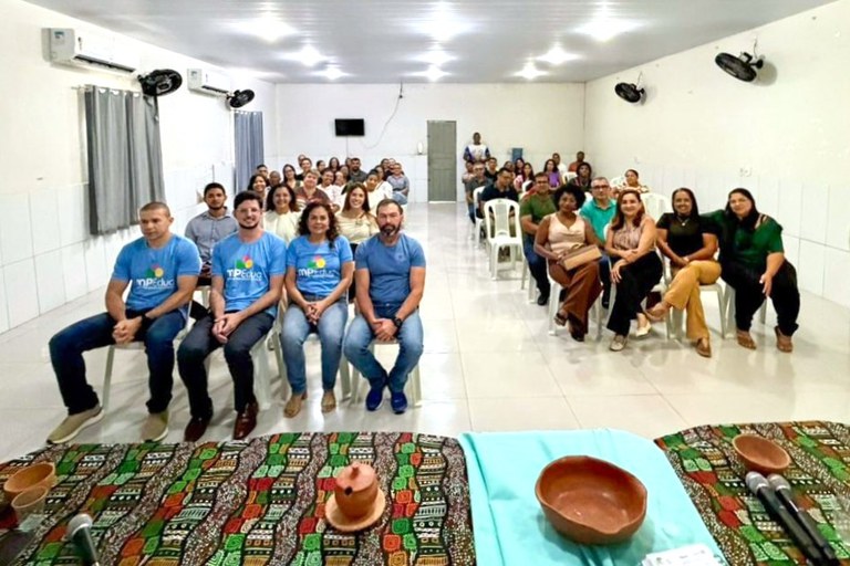 Uma fotografia em plano geral e ângulo plongée (de cima para baixo) mostra uma audiência pública em um amplo salão de piso claro e paredes brancas. Em primeiro plano, vê-se parte de uma mesa decorada com panos estampados e peças artesanais de argila, além de dois microfones. No centro da imagem, o público está organizado em cadeiras brancas de plástico voltadas para a mesa: na primeira fila à esquerda, destacam-se quatro pessoas sentadas lado a lado, sendo que três vestem camisetas azuis com o logotipo colorido do projeto "MPEduc"; atrás delas e em fileiras adjacentes à direita, dezenas de homens e mulheres de idades e vestimentas variadas acompanham o evento. Ao fundo, o espaço amplo exibe luminárias embutidas no teto, aparelhos de ar-condicionado e ventiladores fixados nas paredes laterais, e uma porta fechada ao centro.