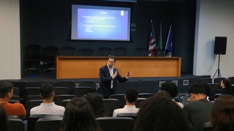 Procurador-chefe do MPF no Maranhão ministra palestra em auditório para estudantes universitários sentados na plateia. Ao fundo, há uma projeção sobre a estrutura da Procuradoria da República no Maranhão.