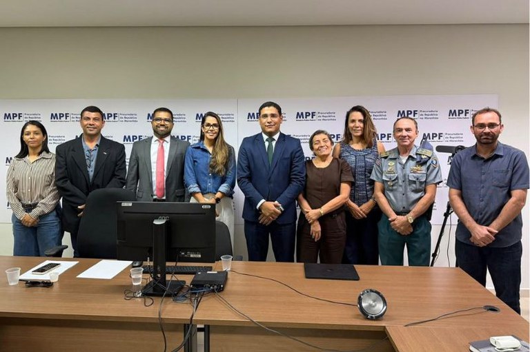 Grupo de nove pessoas, incluindo civis e um militar fardado, posa de pé atrás de uma mesa de reunião em frente a um painel com a sigla e o nome do MPF (Procuradoria da República no Maranhão).