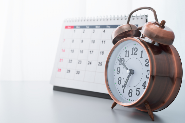 Imagem de um relógio despertador analógico marrom, posicionado sobre uma superfície branca. Ao fundo, há um calendário de mesa desfocado, representando datas e passagem do tempo.