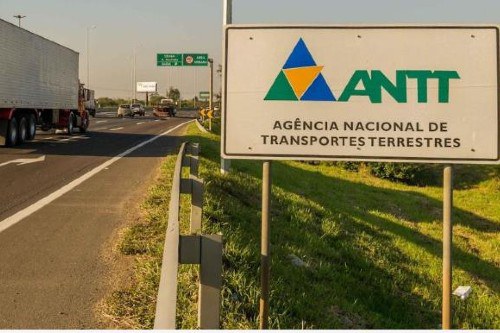 Foto mostra uma placa da ANTT ao lado de uma rodovia federal por onde circulam carros e caminhões
