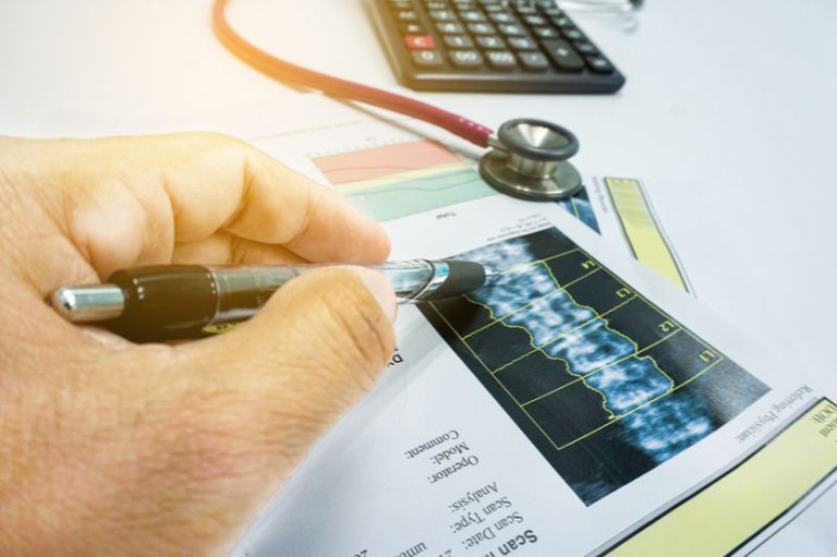 foto de um exame de imagens com o detalhe de uma mão segurando uma caneta apontado para um ponto específico; ao lado um estetoscópio e uma calculadora