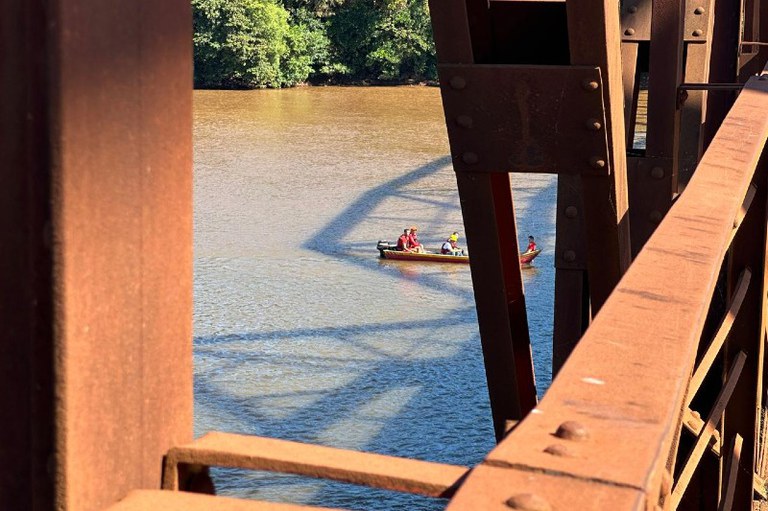 Foto de um detalhe da ponte da revolução mostrando a estrutura e ao fundo, no Rio Grande, uma canoa passando com algumas pessoas dentro