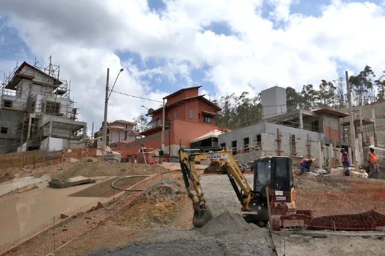 Foto mostra máquina retroescavadeira pegando areia. Ao fundo novas casas em construção