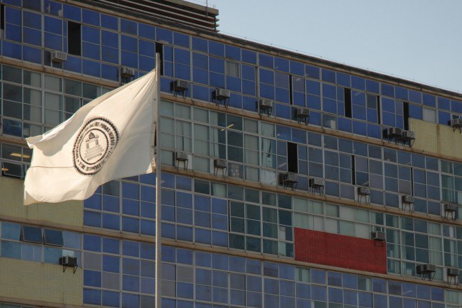 A imagem apresenta o Prédio da Reitoria da Universidade Federal de Minas Gerais (UFMG), destacando sua arquitetura modernista caracterizada por uma fachada de vidro composta por diversos painéis em tons de azul, intercalados com alguns elementos em cinza e um detalhe vibrante em vermelho. Em primeiro plano, à esquerda, uma bandeira branca com o brasão da universidade tremula suavemente, presa a um mastro metálico. A lateral do edifício exibe diversas unidades de ar-condicionado de janela instaladas sob as esquadrias, e o reflexo do céu claro nas janelas confere uma sensação de luminosidade à cena, capturada sob a luz do dia.