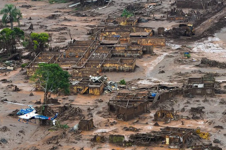 A imagem mostra a destruição causada pelo rompimento de barragem em Mariana (MG), com uma extensa área coberta por lama. Diversas casas estão destruídas ou parcialmente soterradas, e os destroços se espalham por toda a região. Um helicóptero sobrevoa o local.