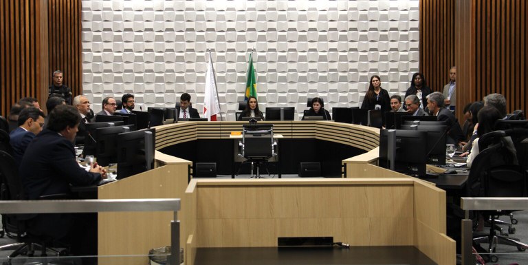 Fotografia de audiência judicial em plenário da Justiça Federal. Diversas pessoas em trajes formais ocupam uma bancada em formato de "U", com as bandeiras de Minas Gerais e do Brasil ao centro.