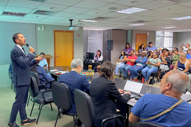 O procurador da República Eduardo Henrique de Almeida Aguiar fala em reunião, realizada em 03/02/2025, entre o MPF, órgãos públicos e pessoas atingidas pelo rompimento da barragem de Fundão, em Minas Gerais. Na foto, podemos ver, à esquerda, o procurador em pé, segurando o microfone, pessoas sentadas à mesa do evento, no centro, e pessoas sentadas no auditório, no lado direito.
