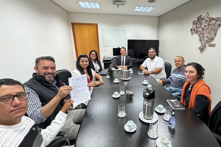 A imagem mostra um grupo de oito pessoas, aparentando ser de diferentes idades e gêneros, sentadas em uma sala de reuniões. Eles estão ao redor de uma grande mesa preta, onde há xícaras de café, um bule, copos e garrafas. Uma pessoa, no lado esquerdo e mais próxima da câmera, segura e mostra um documento. O grupo parece estar engajado em uma conversa ou reunião, com algumas pessoas sorrindo para a câmera. Ao fundo, há uma parede com um grande mapa do Brasil em relevo, e uma porta marrom.