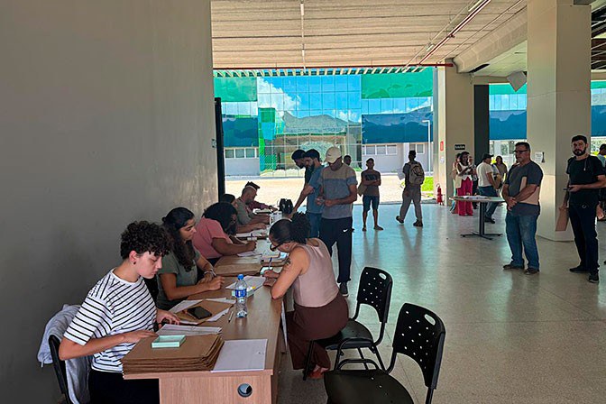 A imagem registra um ambiente interno amplo e bem iluminado, possivelmente o saguão de um prédio público ou educacional, onde ocorre um processo de atendimento ou votação. Em primeiro plano, à esquerda, quatro mulheres estão sentadas atrás de uma longa mesa de madeira, focadas em organizar documentos e preencher formulários manuais. Ao centro e ao fundo, diversas pessoas circulam ou aguardam em pé, algumas carregando pastas, enquanto grandes janelas de vidro ao fundo revelam uma fachada moderna de tons esverdeados e uma paisagem externa ensolarada. A cena transmite uma atmosfera de organização institucional e engajamento comunitário.