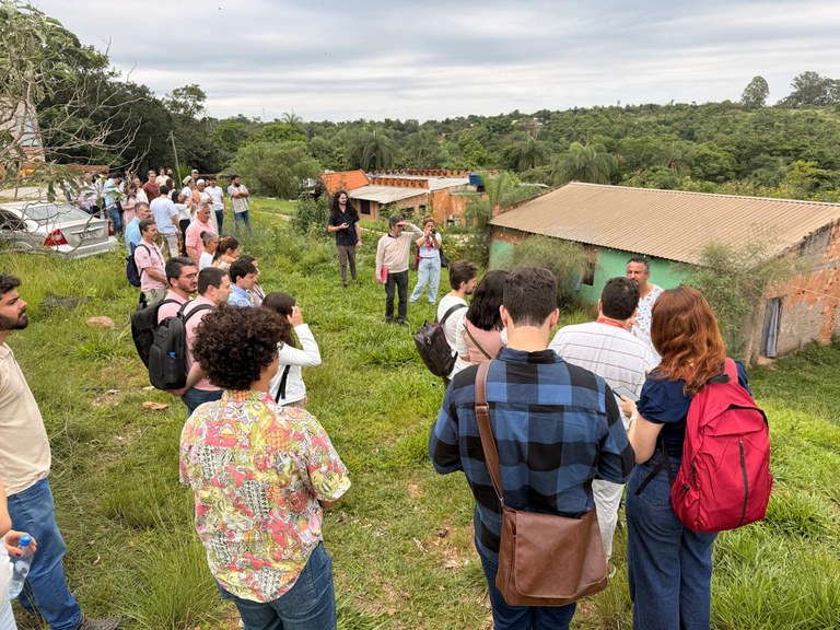 Fotografia ao ar livre mostra um grupo de pessoas reunidas em terreno de vegetação baixa diante de pequenas casas simples. Ao fundo, observam-se árvores e vegetação densa sob céu nublado.