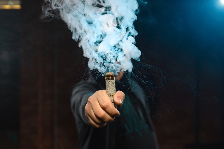 Foto mostra uma pessoa com um vape na mão. A fumaça encobre o rosto