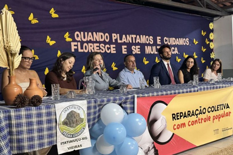 Foto mostra mesa do evento com sete pessoas sentadas, atrás a frase quando plantamos e cuidamos, floresce e se multiplica; na frente um banner com a frase reparação coletiva com controle popular