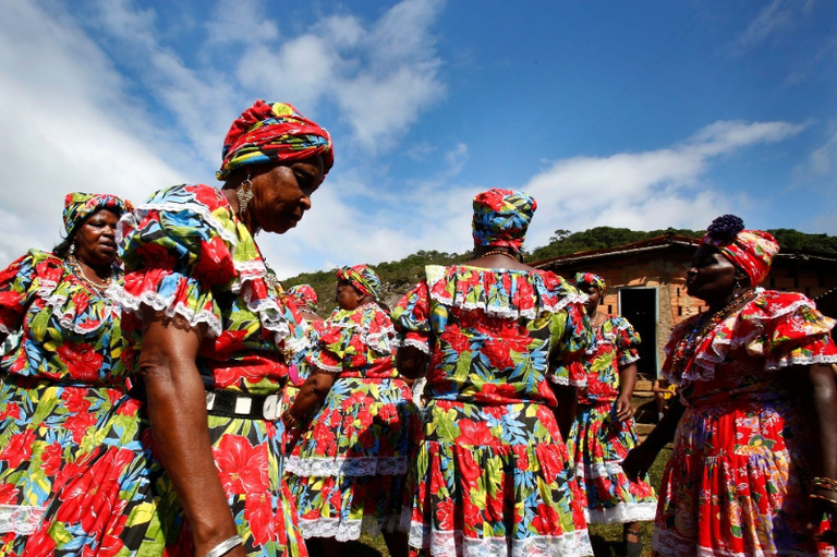 Mulheres negras quilombolas reunidas ao ar livre sob um céu azul com nuvens. Elas vestem trajes tradicionais idênticos: vestidos coloridos com estampas florais vibrantes em vermelho e azul, detalhes em renda branca e turbantes combinando. Ao fundo, vê-se uma construção simples de tijolos e vegetação.