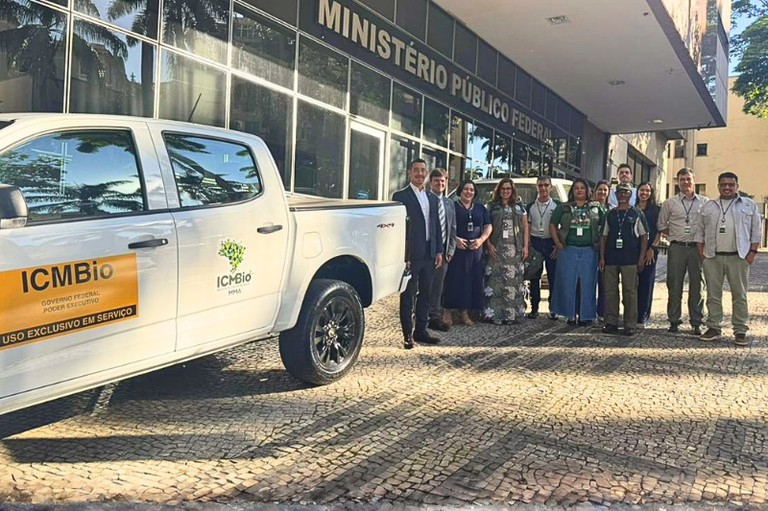 Uma caminhonete branca com adesivos do ICMBio está estacionada em frente a um prédio do Ministério Público Federal. Ao lado do veículo, um grupo de aproximadamente doze pessoas posa para uma foto oficial sobre uma calçada de pedras portuguesas.