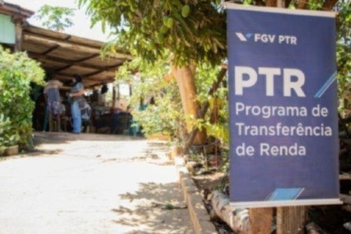 Foto mostra banner onde está escrito Programa de Transferência de Renda e pessoas desfocadas ao fundo