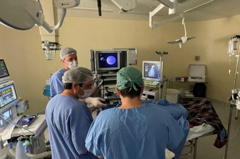 Foto mostra um sala de cirurgia com um paciente na maca, aparelhos e três médicos ao redor