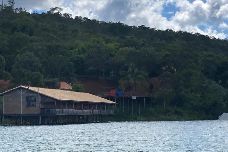 Edificação retangular de madeira construída sobre estacas (palafita) na margem de um reservatório de água, com vegetação densa e um morro ao fundo sob céu ensolarado com nuvens.