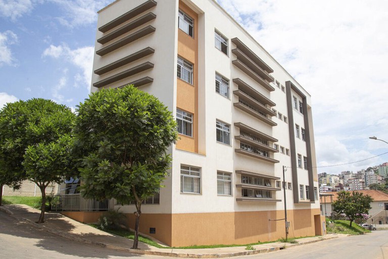 A imagem apresenta o edifício da Unidade de Atendimento Especializado em Saúde (UAES) da Universidade Federal de Viçosa, uma estrutura moderna de múltiplos pavimentos situada em um terreno inclinado. A fachada principal é predominantemente branca com detalhes em tons de ocre na base e em colunas verticais, destacando-se pelas janelas amplas protegidas por brises horizontais cinzas que conferem um ritmo geométrico à arquitetura. No primeiro plano, árvores de copa densa e verde ladeiam a calçada, enquanto ao fundo observa-se o relevo acidentado da cidade com diversas edificações sob um céu claro e parcialmente nublado.