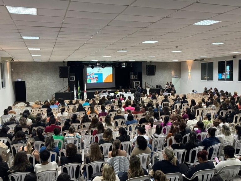 Uma fotografia colorida em plano geral e com ângulo levemente elevado mostra o interior de um amplo auditório lotado durante um evento público. Em primeiro plano e no plano médio, dezenas de fileiras de cadeiras de plástico brancas estão ocupadas por um grande público, majoritariamente composto por mulheres, vistas de costas. Ao fundo, no palco centralizado, há uma mesa comprida com toalha branca onde estão sentadas algumas pessoas de frente para a plateia. Atrás da mesa, uma grande tela de projeção exibe um slide com imagens e textos institucionais coloridos. O auditório possui teto modular branco com várias luminárias embutidas acesas, paredes laterais claras com janelas de vidro à direita e uma parede de fundo cinza ao redor da área do palco.