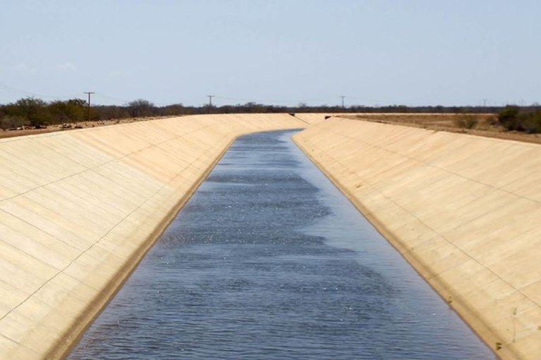 Canal de irrigação revestido de concreto com água fluindo, estendendo-se por uma paisagem rural de vegetação seca sob céu claro.