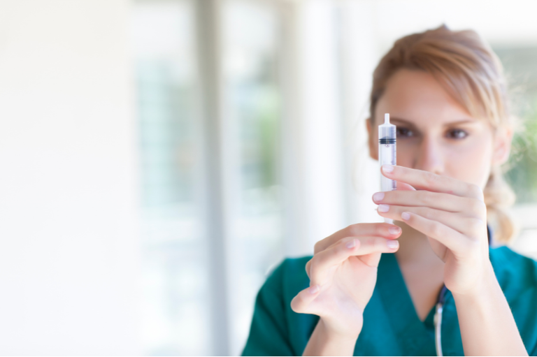 Profissional de saúde usando uniforme verde segura e examina uma seringa transparente, com fundo claro e desfocado.
