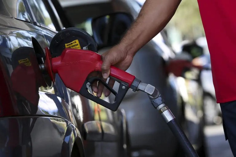 Fotografia em plano aproximado do momento em que um veículo é abastecido em um posto de combustível. No lado esquerdo, vê-se a lateral traseira de um carro cinza-escuro com o bocal do tanque aberto, onde está inserida a ponta metálica do bocal de combustível. No centro, a mão de um frentista, usando camisa vermelha do lado direito, segura o gatilho de uma bico injetor de cor vermelha com mangueira preta. O fundo apresenta outros veículos e o ambiente externo desfocados sob luz natural.