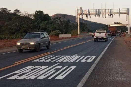 Fotografia em plano geral mostra o trecho de uma rodovia asfaltada com tráfego nos dois sentidos sob a luz do fim de tarde. Na pista da direita, o asfalto novinho traz a sinalização horizontal pintada em letras maiúsculas brancas no chão, lidas de baixo para cima como "PEDÁGIO ELETRÔNICO". Um carro de passeio cinza circula no sentido oposto, à esquerda, enquanto uma picape branca e outros veículos seguem em direção a um pórtico metálico branco de pedágio sem barreiras (sistema Free Flow), equipado com sensores e câmeras, instalado sobre a pista. Ao fundo, a paisagem é composta por vegetação densa e morros ao horizonte.