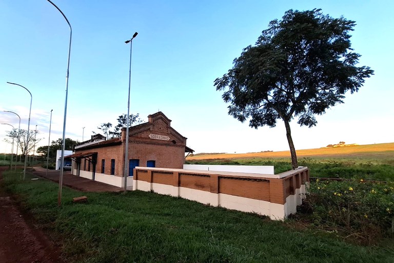 A imagem apresenta a Estação Sobradinho, uma edificação histórica de tijolos aparentes localizada em uma área rural de Uberlândia, Minas Gerais. O prédio possui uma arquitetura ferroviária clássica, com portas e janelas azuis, além de um frontão trabalhado onde se lê o nome da estação. À frente da estrutura, há um muro baixo que combina tons de bege e marrom, acompanhado por um gramado verde e uma árvore solitária de copa densa que se destaca contra o céu claro ao entardecer. A cena é emoldurada por postes de iluminação modernos ao longo de uma estrada de terra, contrastando o patrimônio histórico com a infraestrutura atual, enquanto ao fundo observa-se uma suave colina sob a luz dourada do sol poente.